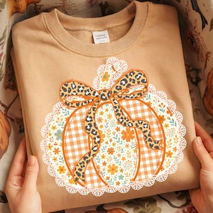 May include: A tan sweatshirt with a pumpkin design. The pumpkin has a gingham pattern, a leopard print bow, and is surrounded by a floral and lace design. The sweatshirt is folded and held by hands.