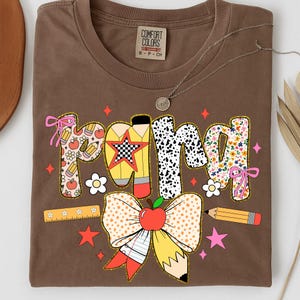 May include: A brown Comfort Colors t-shirt with a colorful "Pom" graphic. The graphic features pencils, apples, flowers, and a large bow with an apple. A silver necklace with a small pendant is also visible.