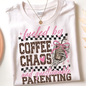 Puede incluir: Camiseta blanca con el texto "fueled by COFFEE CHAOS and questionable PARENTING" en marrón y rosa. Una mano de esqueleto sostiene una taza de café con un lazo rosa. Se ve un collar dorado.