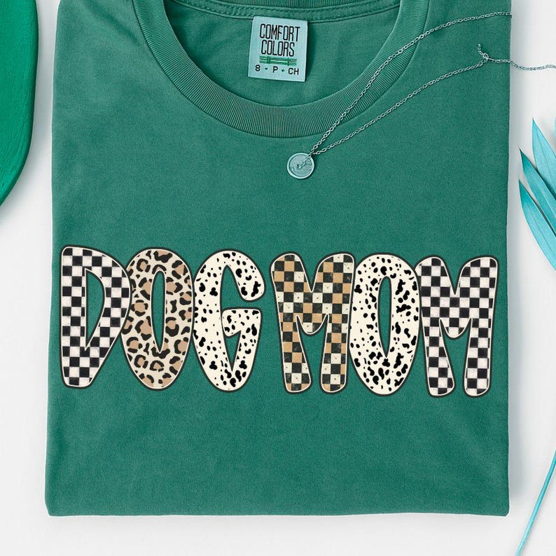 Puede incluir: Una camiseta Comfort Colors verde azulado con el texto "DOG MOM" en una mezcla de estampados, incluyendo estampado de leopardo y dise&ntilde;os a cuadros. Se ve un collar plateado con un peque&ntilde;o colgante. La camiseta tiene un cuello redondo cl&aacute;sico.
