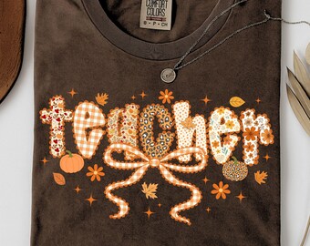 May include: A brown t-shirt with the word "teacher" in a decorative font. The letters are filled with floral, plaid, and leopard print patterns. The design includes pumpkins, leaves, and a bow. A silver necklace is draped across the shirt.
