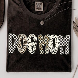 Puede incluir: Una camiseta marr&oacute;n oscuro con las palabras "DOG MOM" en una fuente audaz y juguetona. Las letras presentan una mezcla de patrones, incluyendo estampado de leopardo, cuadros y lunares d&aacute;lmata. Se ve un collar plateado con un peque&ntilde;o colgante.