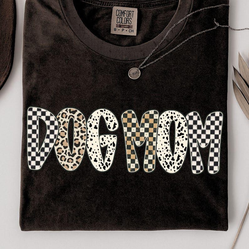 Puede incluir: Una camiseta marr&oacute;n oscuro con las palabras "DOG MOM" en una fuente audaz y juguetona. Las letras presentan una mezcla de patrones, incluyendo estampado de leopardo, cuadros y lunares d&aacute;lmata. Se ve un collar plateado con un peque&ntilde;o colgante.