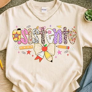 Puede incluir: Camiseta color crema con la palabra "Assistant" en un diseño colorido y estampado. El diseño incluye un lazo con una manzana, un lápiz y otros elementos temáticos escolares. La camiseta tiene cuello redondo y mangas cortas.