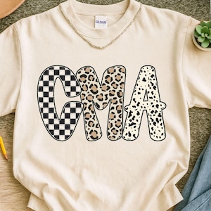 May include: Cream-colored t-shirt with the word "CMA" in a bold, patterned design. The letter "C" features a black and white checkerboard pattern, "M" has a leopard print, and "A" has a dalmatian print. The shirt has a crew neck and short sleeves.