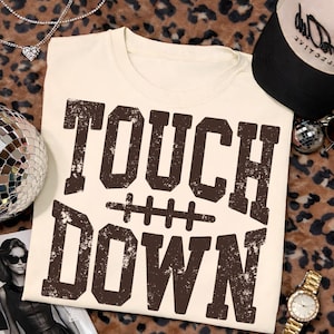 May include: A cream-colored t-shirt featuring the dark brown text "TOUCH DOWN" in a distressed, block-style font. Between the two words is a graphic of a football lace. The shirt has a crew neckline and short sleeves, presented in a flat layout.