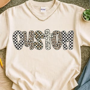 Könnte beinhalten: Cremefarbenes T-Shirt mit dem Wort "CUSTOM" in einem auffälligen, mehrfach gemusterten Design. Die Buchstaben weisen eine Mischung aus schwarz-weiß karierten, Leopardenmuster- und gesprenkelten Mustern auf. Eine goldene Halskette ist am Ausschnitt zu sehen.