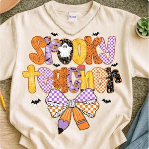 May include: Cream-colored sweatshirt with the words "Spooky Teacher" in a colorful, Halloween-themed design. The design includes bats, ghosts, and a pencil-shaped bow. The sweatshirt has a crew neck and the brand name "Gildan" is visible.