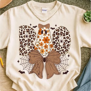 May include: Cream-colored sweatshirt with a Halloween-themed design. The word "BOO" is spelled out with leopard print letters. A ghost with orange flowers is in the center, and a brown bow and spiderweb are at the bottom.