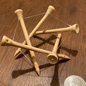 May include: A geometric structure made of several wooden golf tees, connected by thin, clear string. The tees are light brown, with a rounded top and a pointed end. A US quarter coin is next to the structure on a dark wood surface.