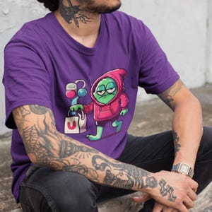 Könnte beinhalten: Lilafarbenes T-Shirt mit einem Cartoon-Frosch, der eine rote Kapuze trägt und eine Einkaufstasche hält. Der Frosch hat grüne Haut, schwarze Augen und einen weißen Mund. Die Einkaufstasche ist weiß mit einem roten "U" darauf.