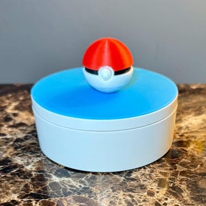 May include: A white and blue round container with a red and white ball-shaped lid. The lid is a stylized representation of a Pokeball. The container sits on a brown and gray surface.