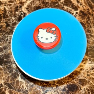 May include: A blue and white Hello Kitty themed lid. The lid has a red center with a white Hello Kitty face and a red bow. The lid is round and appears to be made of plastic.