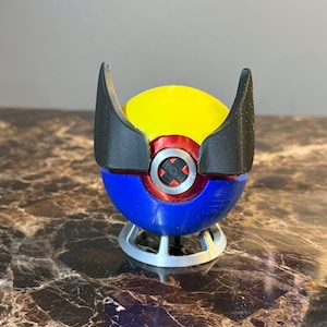 Pode incluir: Uma Pokébola impressa em 3D com um design amarelo, vermelho e azul. A Pokébola tem asas pretas no topo e uma base prateada.
