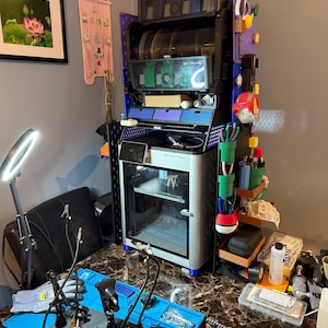 May include: A multi-tiered, black and blue storage unit with clear, circular containers. A 3D printer sits below, with a blue mat and tools in the foreground. A painting and a pink banner are on the wall.