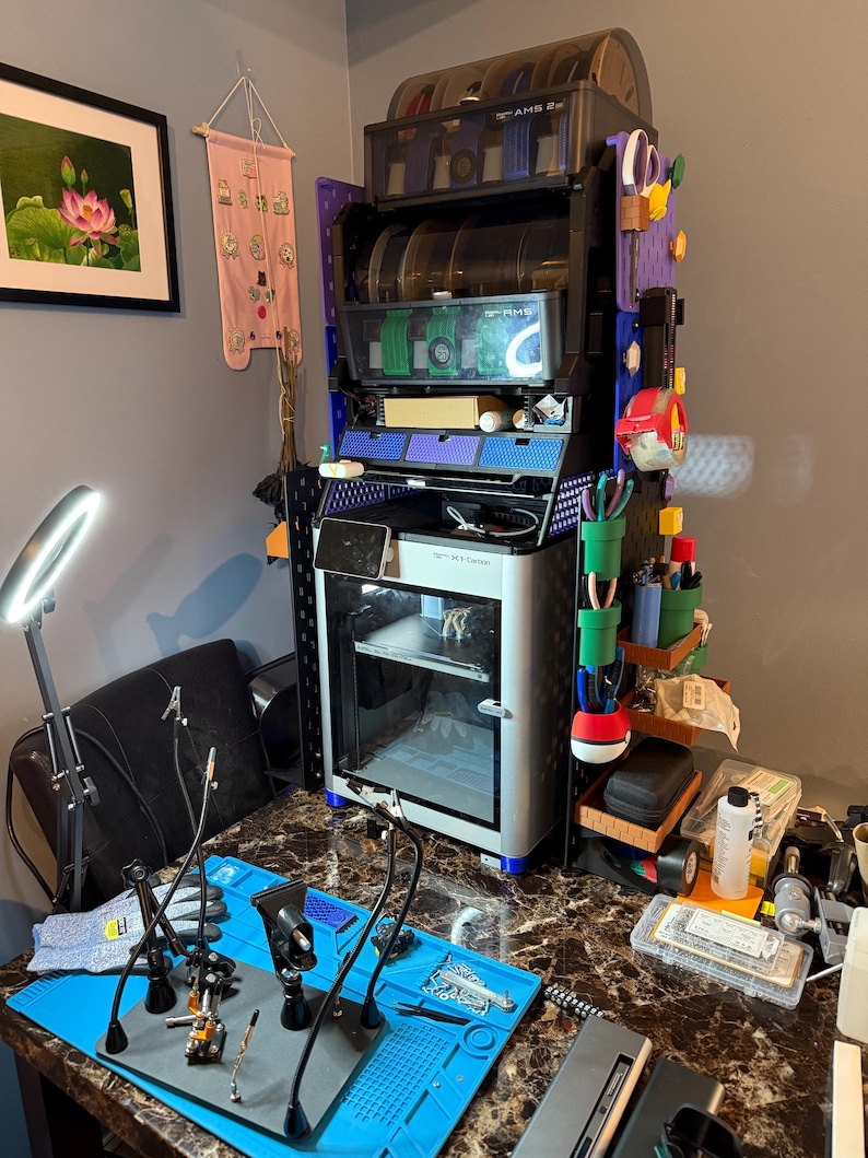 May include: A multi-tiered, black and blue storage unit with clear, circular containers. A 3D printer sits below, with a blue mat and tools in the foreground. A painting and a pink banner are on the wall.