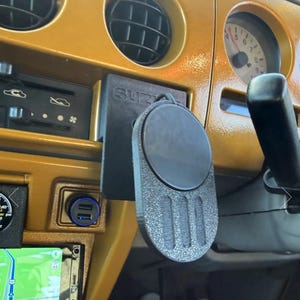Puede incluir: Soporte negro para teléfono con el texto "SUZUKI" en un salpicadero dorado. El soporte tiene un área magnética circular y una base rectangular texturizada. Un puerto de carga USB es visible debajo.