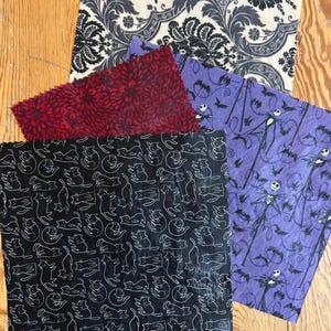 May include: A set of four square fabric wraps featuring distinct patterns: a black and white damask print, a deep red floral design, a black fabric with white line-art cats, and a purple fabric with a repeating character and bat motif.