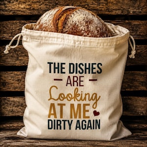May include: A cream-colored fabric drawstring bag containing a round loaf of crusty brown bread. The bag features dark teal and gold text that reads: THE DISHES ARE Looking AT ME DIRTY AGAIN, with a small red heart icon next to the word ME.