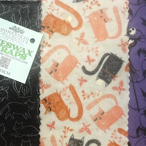 May include: Three rectangular beeswax wraps featuring distinct patterns: a black fabric with white cat outlines, a cream fabric with orange and black cats, and a purple fabric with bat and skeleton character prints. A square label reads Homestead Collective.