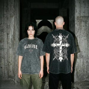May include: Two people wearing dark gray t-shirts. One shirt has the word "saint" in white gothic lettering. The other shirt has a white cross design on the back. Both shirts have a distressed, vintage look.