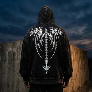 May include: A black hooded sweatshirt featuring a white, stylized spine and wing design on the back. The design is detailed and graphic, with a distressed, vintage appearance. The hoodie has a drawstring and a front pocket.