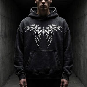 May include: Dark grey hoodie with a white graphic design. The design is a symmetrical wing-like pattern. The hoodie has a kangaroo pocket and a drawstring hood. The fabric has a washed effect.