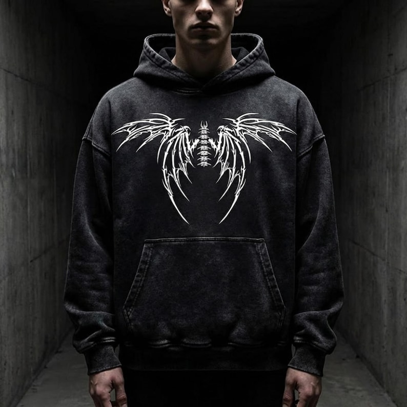 May include: Dark grey hoodie with a white graphic design. The design is a symmetrical wing-like pattern. The hoodie has a kangaroo pocket and a drawstring hood. The fabric has a washed effect.