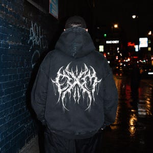 May include: A black hooded sweatshirt featuring a large, white, intricate, symmetrical graphic print on the back. The design consists of sharp, elongated, and jagged lines that form a complex, abstract pattern across the center of the back.