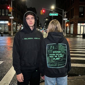 Könnte beinhalten: Zwei Personen tragen schwarze Kapuzenpullover mit grünem Text. Auf einem Kapuzenpullover steht vorne '> RUN_EXECUTE: CHAOS.exe'. Der andere Kapuzenpullover hat hinten eine Grafik mit der Aufschrift 'SYSTEM SYSTEM FAILURE // HACK THE PLANET'.