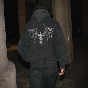 May include: A charcoal black hooded sweatshirt featuring a large, intricate white graphic on the back. The design depicts a stylized, sharp-edged cross with wing-like extensions that resemble thorny, organic branches or metallic filigree.