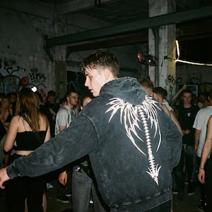 May include: A dark grey hoodie featuring a white spine and wing design on the back. The hoodie is worn by a person in a crowded, dimly lit room with graffiti on the walls. The hoodie has a vintage, worn appearance.