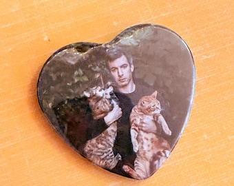 May include: A heart-shaped pin featuring a photographic print of a person with short dark hair wearing a black shirt, holding a brown tabby cat in one arm and an orange tabby cat in the other arm against a dark, leafy green background.