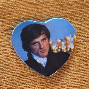 Puede incluir: Espejo en forma de corazón con un retrato de un hombre con vestimenta de época. La imagen tiene un tinte azul y una superficie reflectante. El hombre lleva un abrigo oscuro y una camisa blanca con volantes.