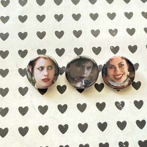 May include: Three round pinback buttons featuring images of women. The buttons have a glossy finish and are set against a white background with a pattern of black hearts. The women have various expressions.