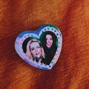 May include: A heart-shaped button featuring two women. The button has a blue and purple gradient background with a white lace-like border. The women are smiling and looking at the camera. The button is on a brown, textured surface.