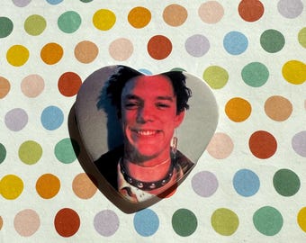 May include: Heart-shaped button featuring a smiling person with dark hair, wearing a black studded choker and earrings. The background is a colorful polka dot pattern with various pastel and bright colors.