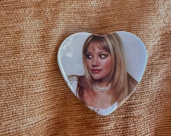 May include: A heart-shaped pin featuring a photographic portrait of a blonde person with shoulder-length hair, wearing a multi-strand pearl necklace and a white top. The image is clear and centered within the heart-shaped frame.