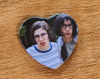 May include: A heart-shaped pin featuring a photographic print of two individuals with dark hair and glasses. One person wears a blue and white striped shirt, while the other wears a grey top with a floral pattern and a dark jacket with a fur-trimmed collar.