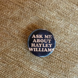 May include: A circular pinback button featuring a dark blue background with the white text "ASK ME ABOUT HAYLEY WILLIAMS" printed in a bold, sans-serif font. The button has a glossy finish and a smooth, rounded edge, presenting a clear message.