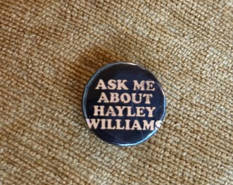 May include: A circular pinback button featuring a dark blue background with the white text "ASK ME ABOUT HAYLEY WILLIAMS" printed in a bold, sans-serif font. The button has a glossy finish and a smooth, rounded edge, presenting a clear message.