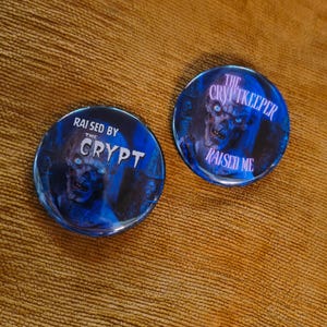 Peut inclure: Deux boutons ronds bleus sur le thème de l'horreur. L'un porte l'inscription "RAISED BY THE CRYPT" avec un graphisme de zombie. L'autre porte l'inscription "THE CRYPTKEEPER RAISED ME" avec un graphisme de zombie similaire. Les deux boutons ont une finition brillante.
