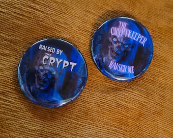 May include: Two round, blue buttons with a horror theme. One button reads "RAISED BY THE CRYPT" with a zombie graphic. The other button reads "THE CRYPTKEEPER RAISED ME" with a similar zombie graphic. Both buttons have a glossy finish.