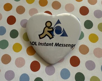 May include: A heart-shaped pinback button with a white background. The design features the tan-colored running person icon and a blue triangle logo of AOL Instant Messenger, with the black text AOL Instant Messenger printed below the icons.