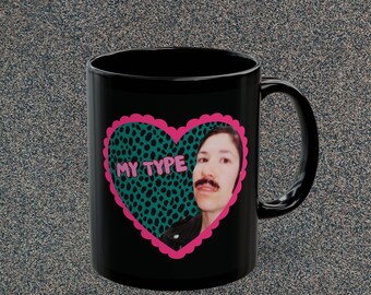 May include: Black ceramic mug featuring a pink heart with a teal leopard print design. The heart contains a photo of a person with a mustache and the words "MY TYPE". The mug has a black handle.