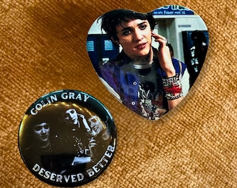 May include: A heart-shaped pin featuring a person with dark hair and a blue jacket, and a circular black pin with a monochrome photograph of three people and the white text "COLIN GRAY DESERVED BETTER" printed along the top and bottom edges.