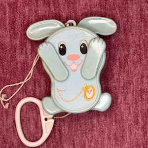 Puede incluir: Un juguete para tirar con forma de conejito azul claro con una nariz rosa y una boca sonriente. El juguete tiene ojos negros grandes y un diseño de reloj de bolsillo dorado. Una cuerda blanca y un asa blanca están unidas.