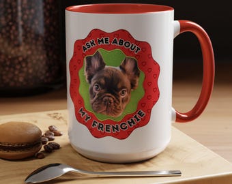 May include: A white ceramic mug with a red interior and handle. The front features a red scalloped border with small heart patterns, a green circular center, and a brown French Bulldog face. The text reads: ASK ME ABOUT MY FRENCHIE.