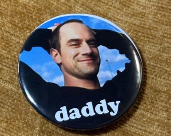 May include: A circular pin-back button featuring a photographic portrait of a person with short dark hair wearing a black shirt against a bright blue sky background. The word "daddy" is printed in bold white lowercase letters at the bottom.