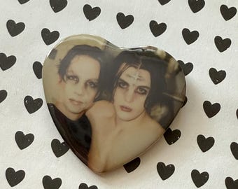 May include: Heart-shaped pin featuring a vintage photograph of two people with dark makeup and hair. The pin is set against a white background with a pattern of black hearts.
